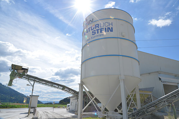 Silo mit Aufschrift 'Natürlich Stein aus Kärnten' auf dem Werksgelände mit Förderband